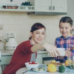 Smiling mother making selfie portrait together with young cute daughter cooking breakfast at home in kitchen. Family, cook, and people concept